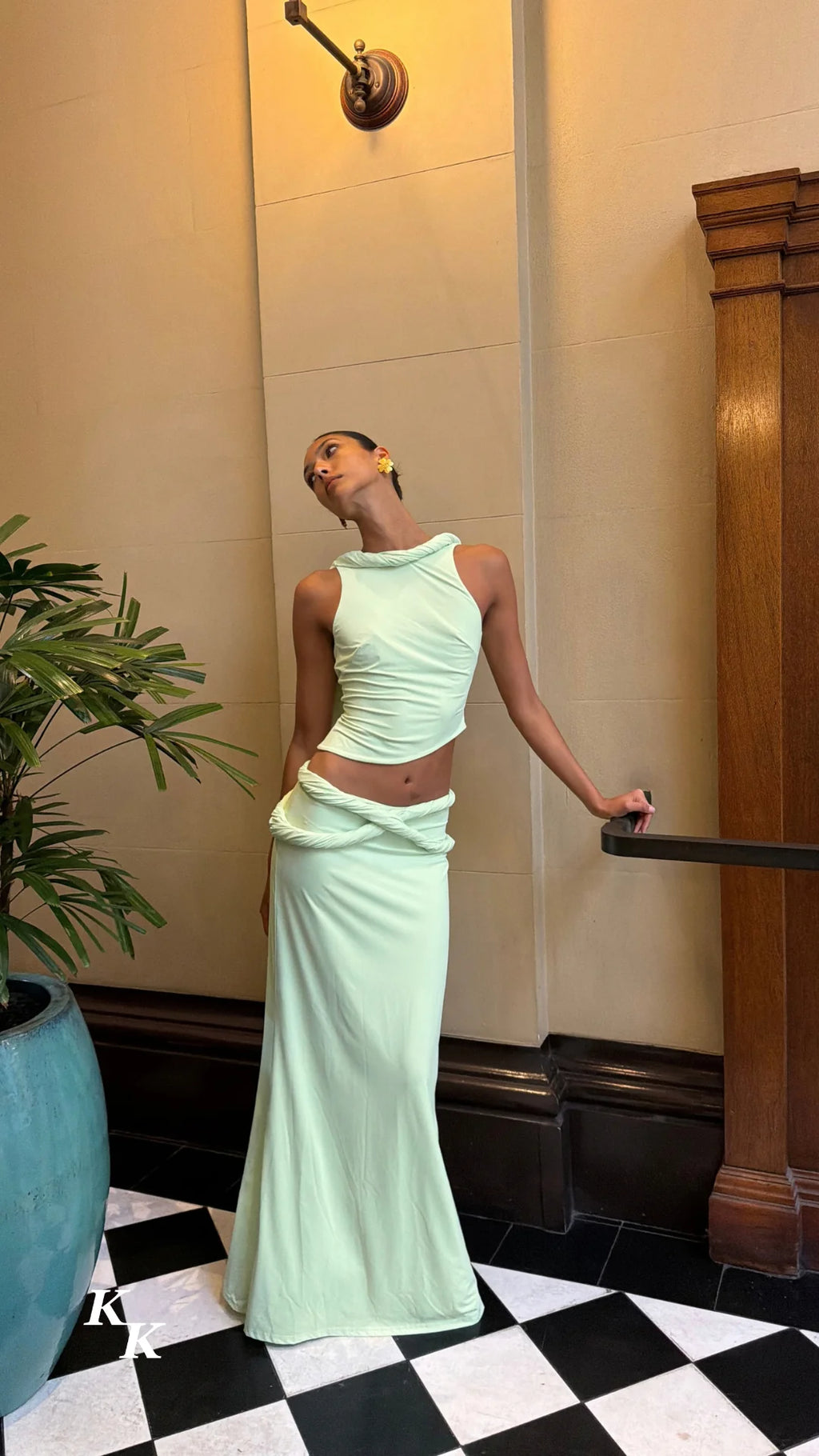 Model wearing the Koster Kulture Athena Set in soft lime green, featuring a high-neck sleeveless top with twisted detailing and a matching low-waisted maxi skirt with a draped waistband.