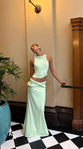 Model wearing the Koster Kulture Athena Set in soft lime green, featuring a high-neck sleeveless top with twisted detailing and a matching low-waisted maxi skirt with a draped waistband.
