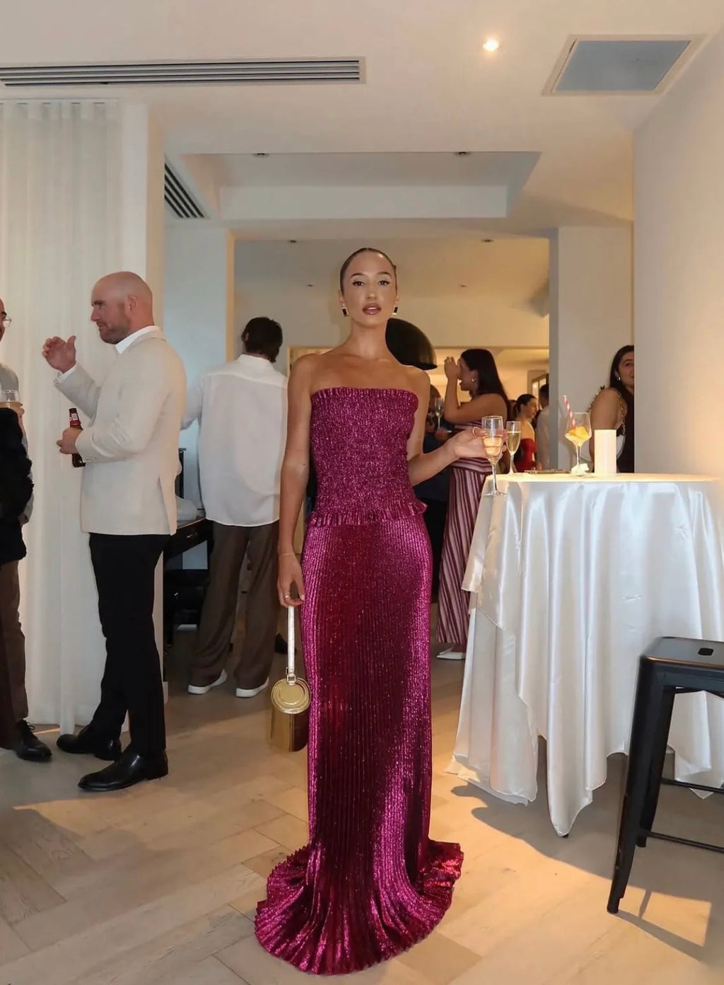 Woman wearing the L’IDÉE Maya Gown in Wine, featuring a strapless ruched bodice, peplum waist and flowing floor-length pleated skirt, styled with a gold handbag at a formal event.