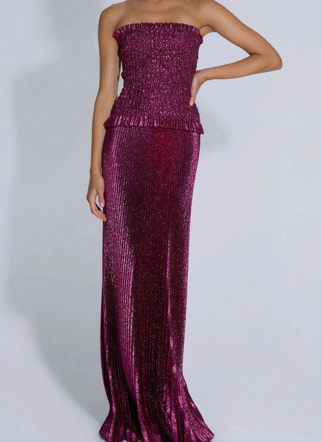 Model wearing the L’IDÉE Maya Gown in Wine, featuring shimmering metallic pleated fabric, a strapless elasticated ruched bodice, delicate peplum waist and flowing floor-length skirt against a neutral studio background.