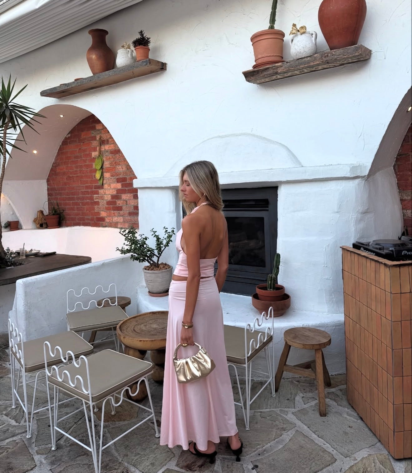 Woman in a pink dress standing in a rustic outdoor setting with white walls, stone floor, and potted plants.