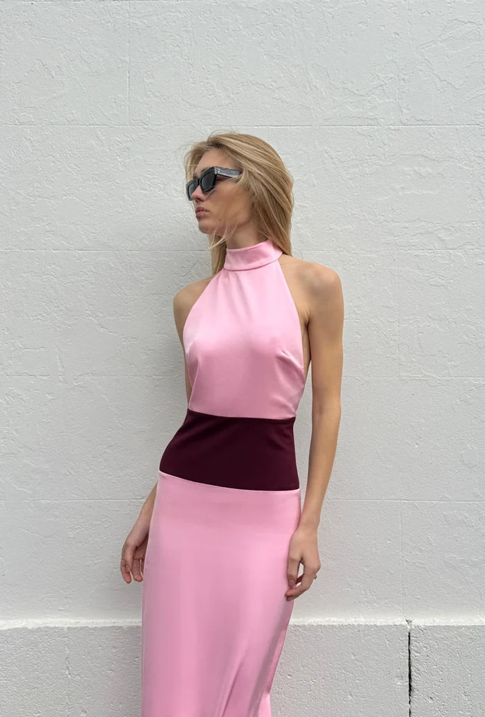 Model wearing the Venetia Maxi Dress in Wine Pink, featuring a soft pink satin halter neckline, contrasting deep burgundy waistband and sleek fitted silhouette, styled with black sunglasses against a white textured wall.
