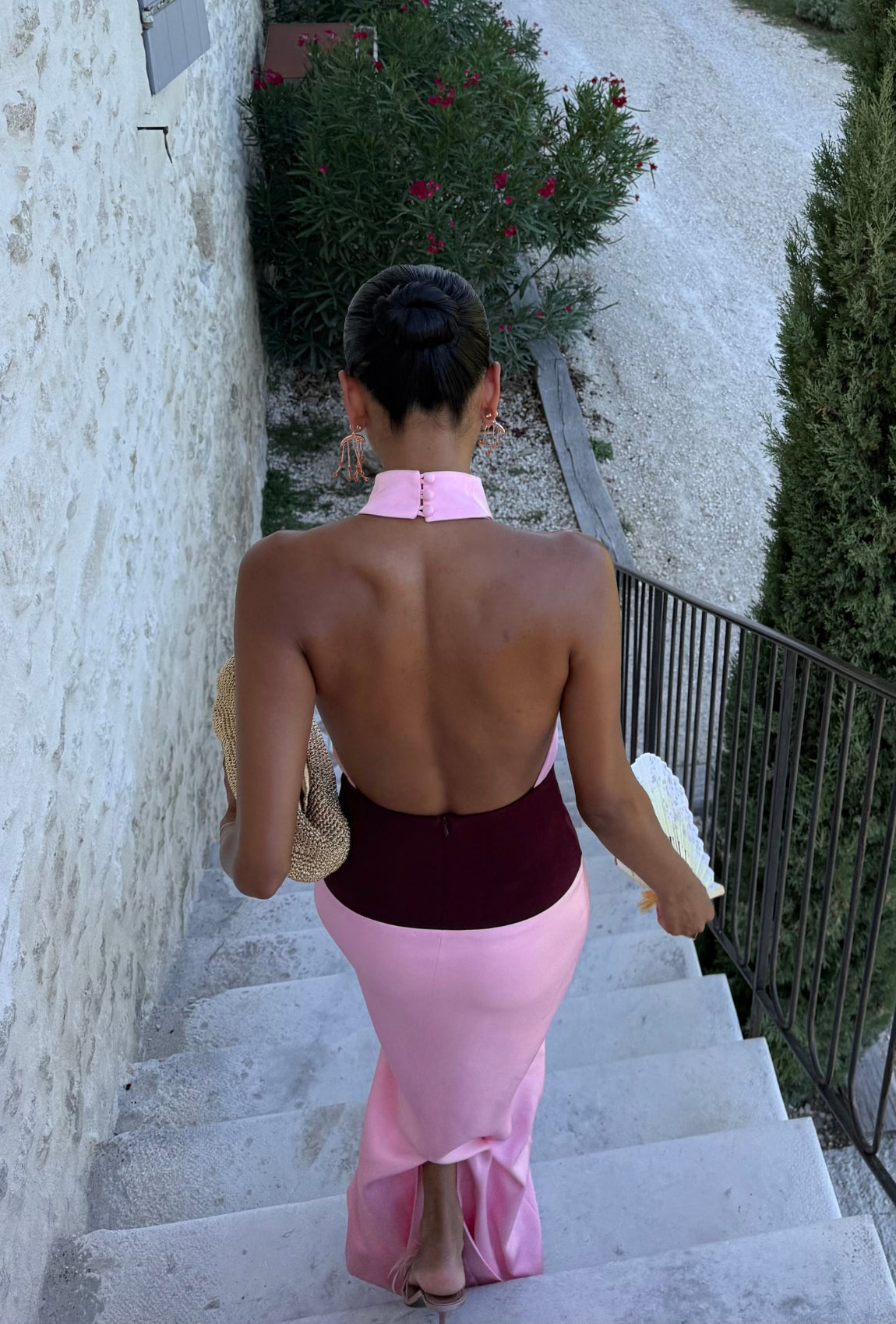 Back view of a model wearing the Venetia Maxi Dress in Wine Pink, featuring a backless halter neckline with fabric-covered button closure, contrasting burgundy waistband and flowing pink maxi skirt, styled with statement earrings and a woven bag on outdoor stone stairs.