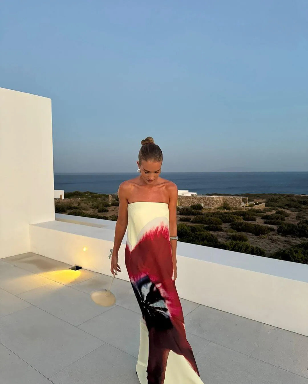 Model wearing a strapless maxi dress featuring a cream base with bold red, burgundy and black abstract floral print, styled with statement earrings and a cream handbag on a modern coastal terrace overlooking the ocean at sunset.