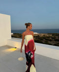 Model wearing a strapless maxi dress featuring a cream base with bold red, burgundy and black abstract floral print, styled with statement earrings and a cream handbag on a modern coastal terrace overlooking the ocean at sunset.