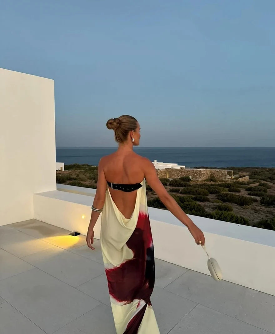 Back view of a model wearing a strapless maxi dress featuring a cream, burgundy and black abstract print, a dramatic draped cowl back and adjustable black leather belt strap, styled with silver accessories on a coastal terrace overlooking the ocean.