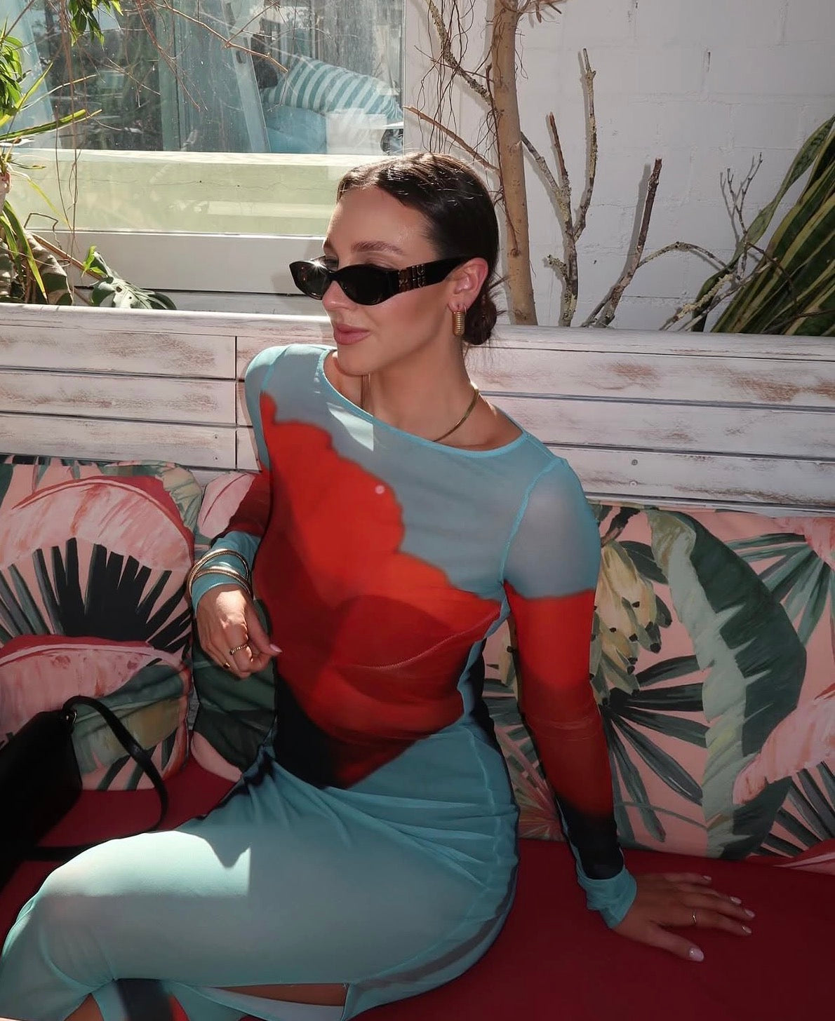 Woman sitting on a colourful couch wearing a red and blue outfit with sunglasses. - @alesha.beggs