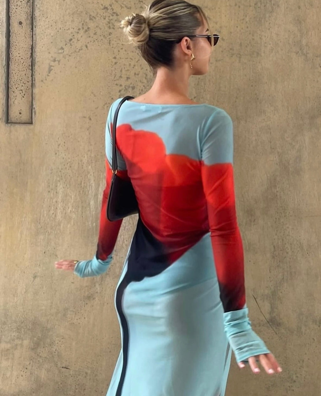 Woman wearing a red and blue dress with a black handbag against a beige wall.