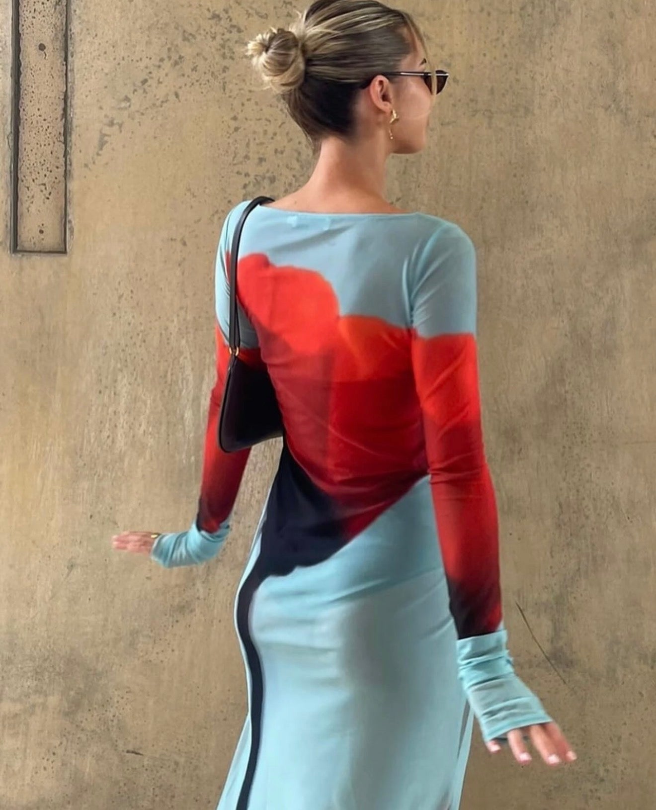 Woman wearing a red and blue dress with a black handbag against a beige wall.