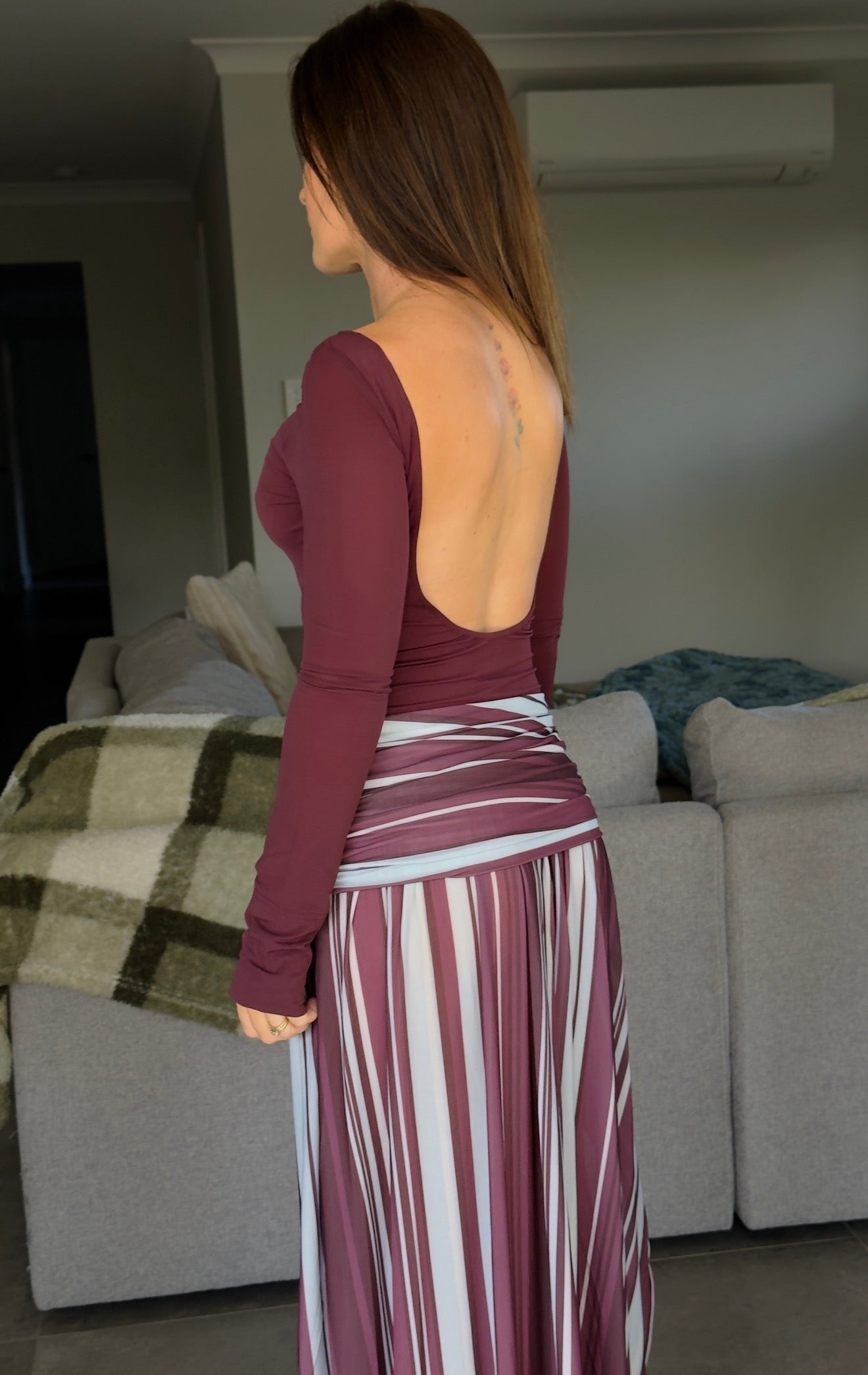 Woman wearing a burgundy open-back top and striped skirt in a living room.
