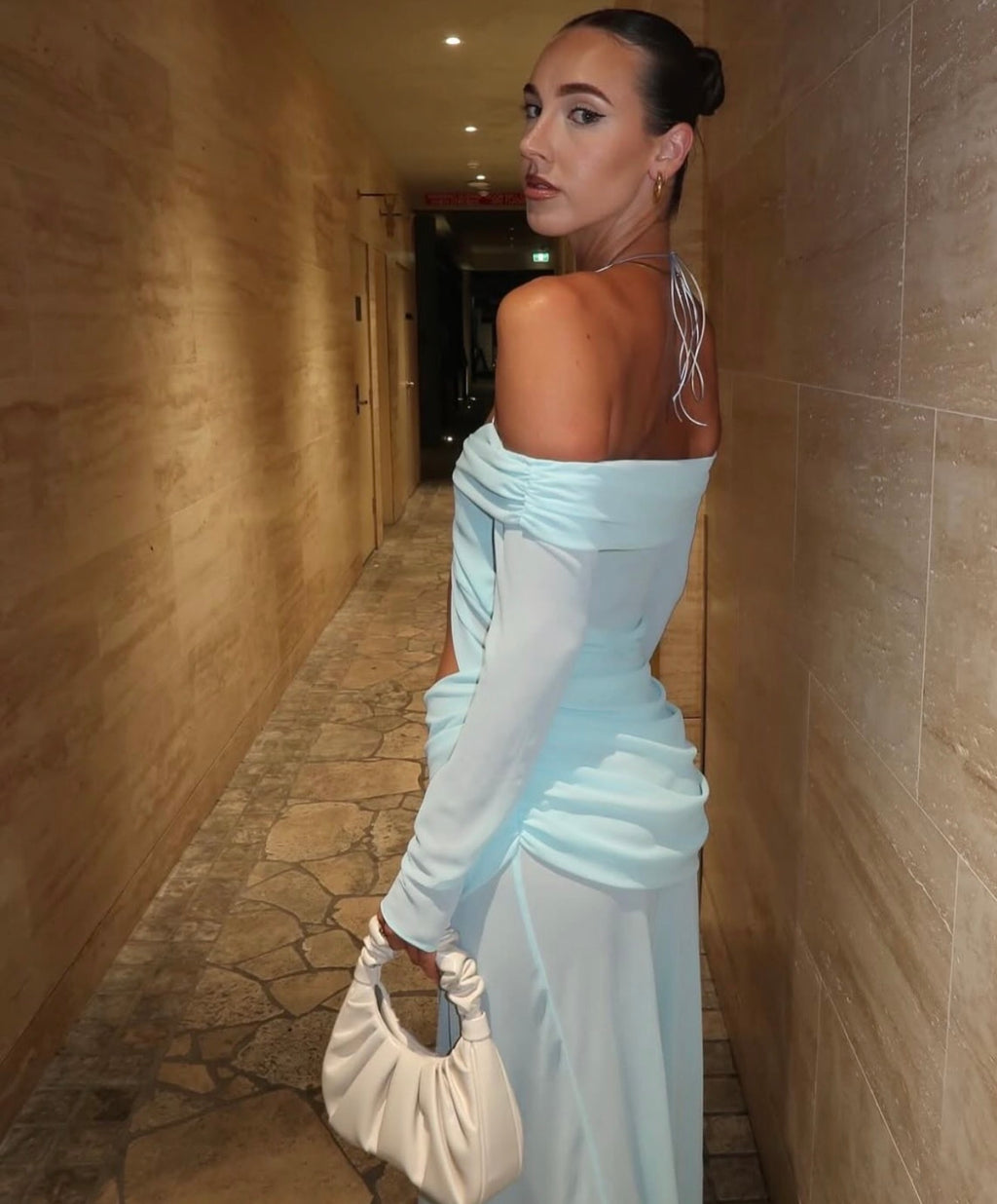 Woman in a light blue off-shoulder dress holding a beige handbag in a hallway.