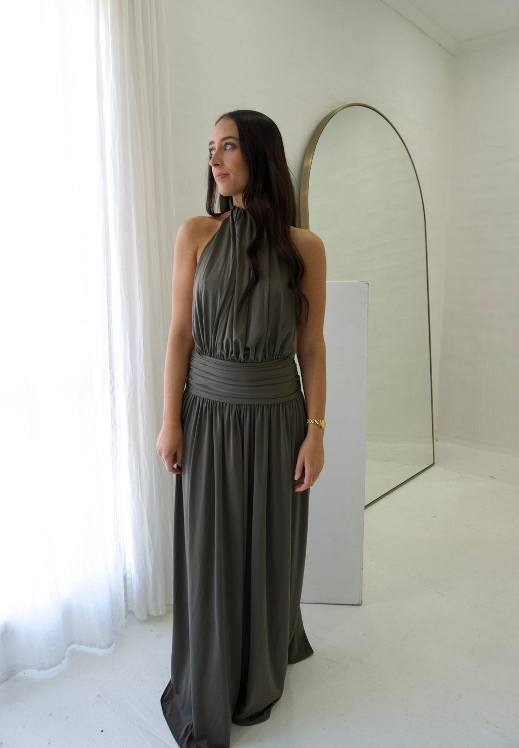 Woman in a long gray dress standing in a minimalistic room with a mirror.