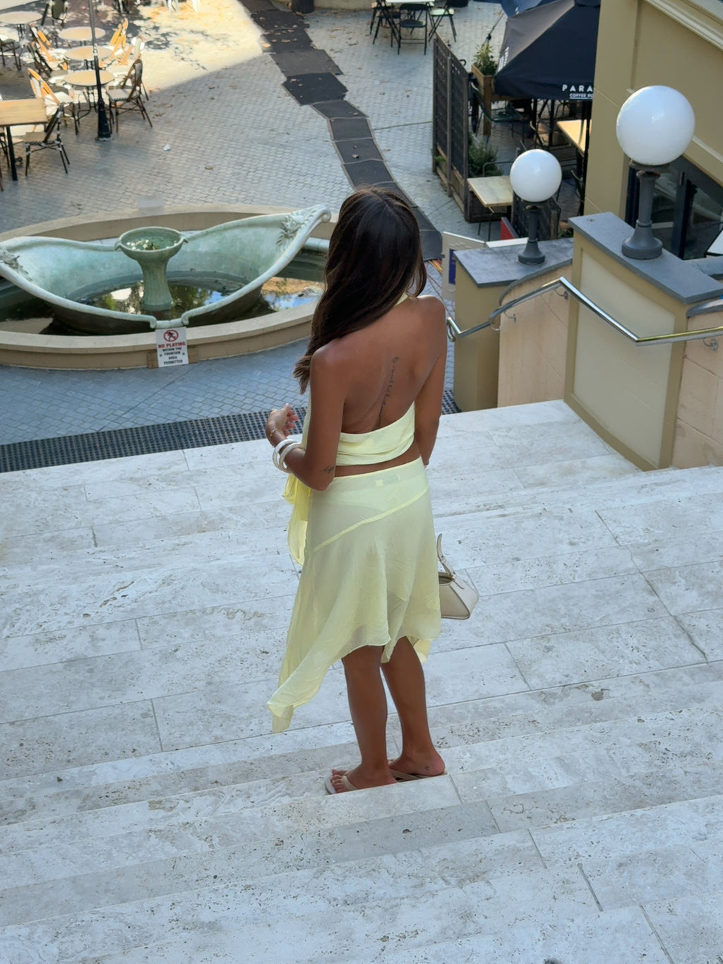 Woman in a yellow dress standing on a stone staircase with a fountain and outdoor seating area in the background.