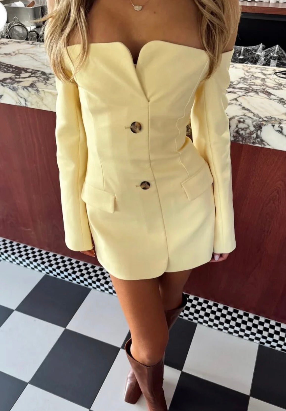 A woman wearing a pale yellow off-the-shoulder mini blazer with double-breasted buttons and brown ankle boots, standing in front of a marble counter and checkered floor.