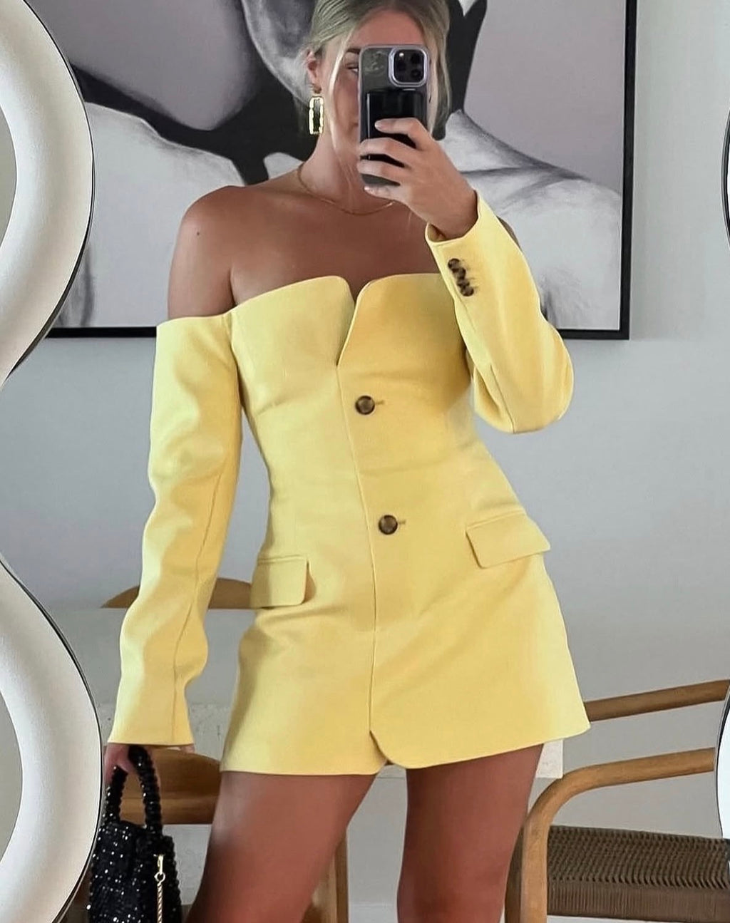 A woman takes a mirror selfie wearing a bright yellow off-the-shoulder blazer-style mini dress with long sleeves and front buttons. The setting appears to be an indoor room with a framed black-and-white wall print, a chair, and a handbag visible.