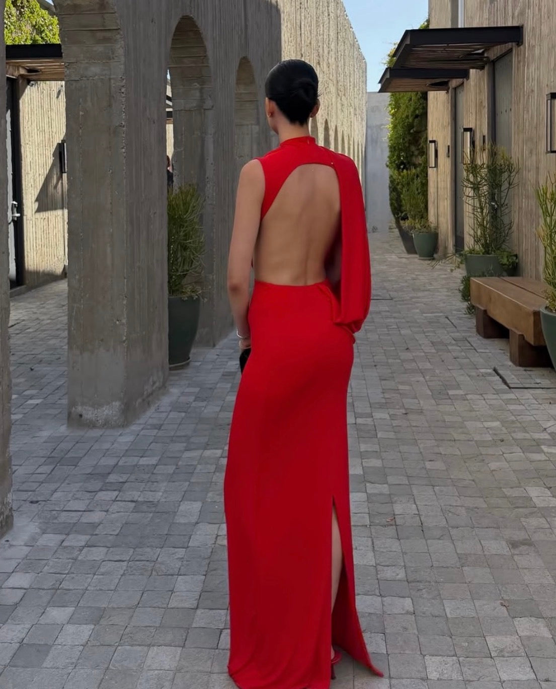 A woman stands in a paved outdoor walkway wearing a bright red sleeveless maxi dress with a dramatic open back and a high slit, photographed from behind.
