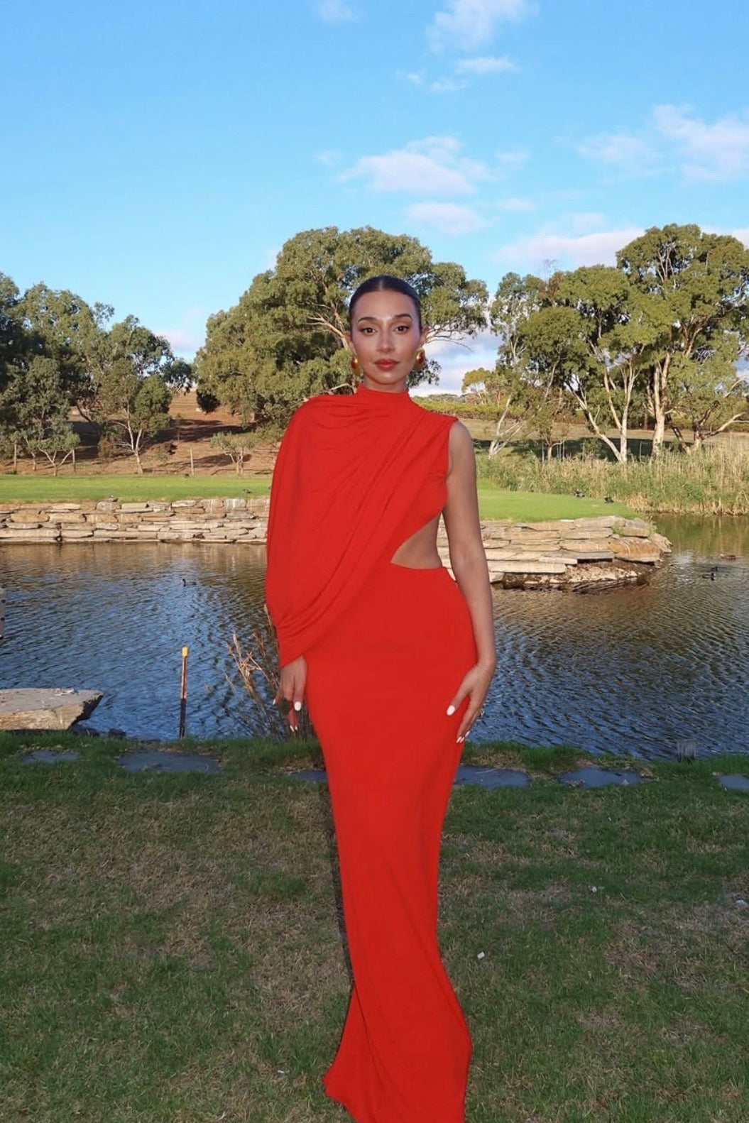 A woman wearing a bright red draped maxi dress with a side cutout, standing outdoors in front of a pond with trees and grass in the background.