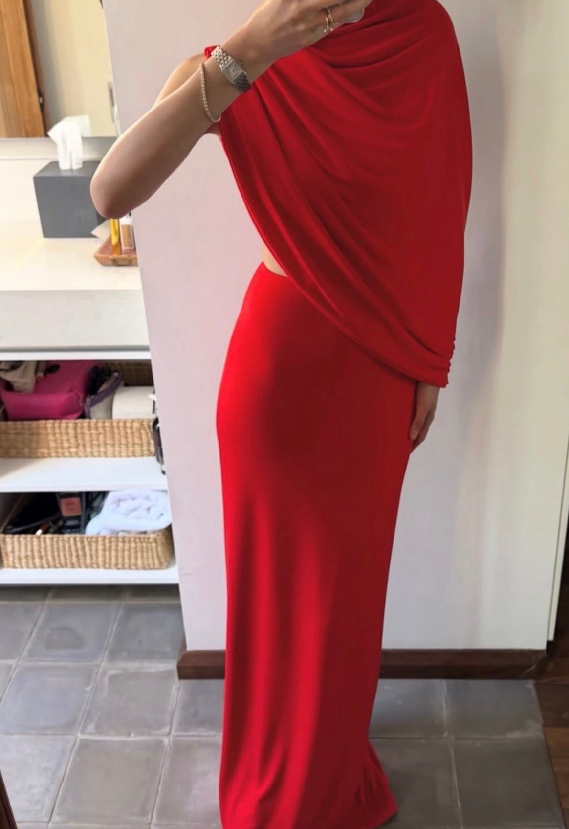 A woman modeling a long, draped red maxi dress in an indoor setting with a white wall and tiled floor visible.