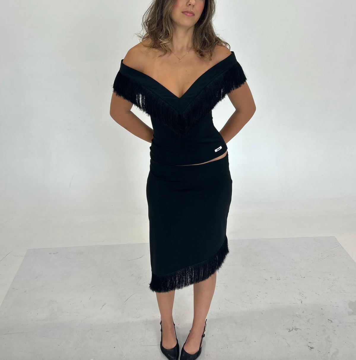 Model wearing the Nalissa Luna Set in black, featuring an off-the-shoulder V-neck top with statement fringe detailing and a matching low-rise midaxi skirt with an asymmetrical fringed hem.