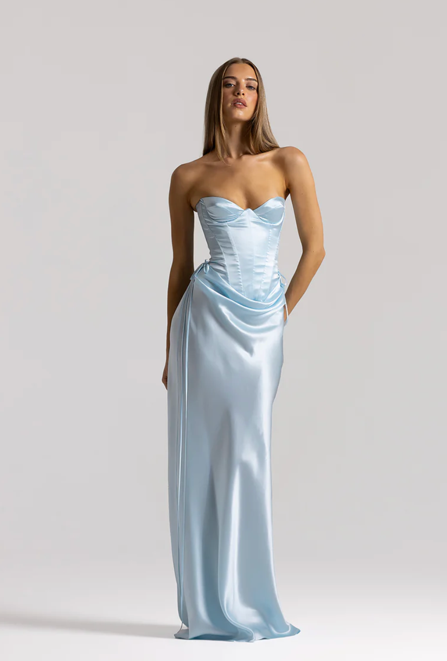 A woman modeling a strapless light blue satin gown with a structured corset-style bodice and draped skirt, photographed against a clean light gray studio background.