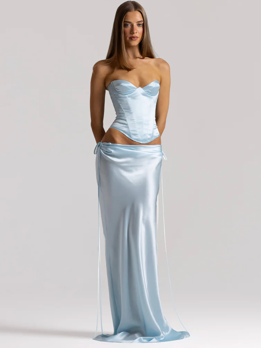 A model wearing a light blue satin two-piece set consisting of a strapless corset-style top and a matching satin wrap skirt with long side ties, photographed against a plain light gray studio background.