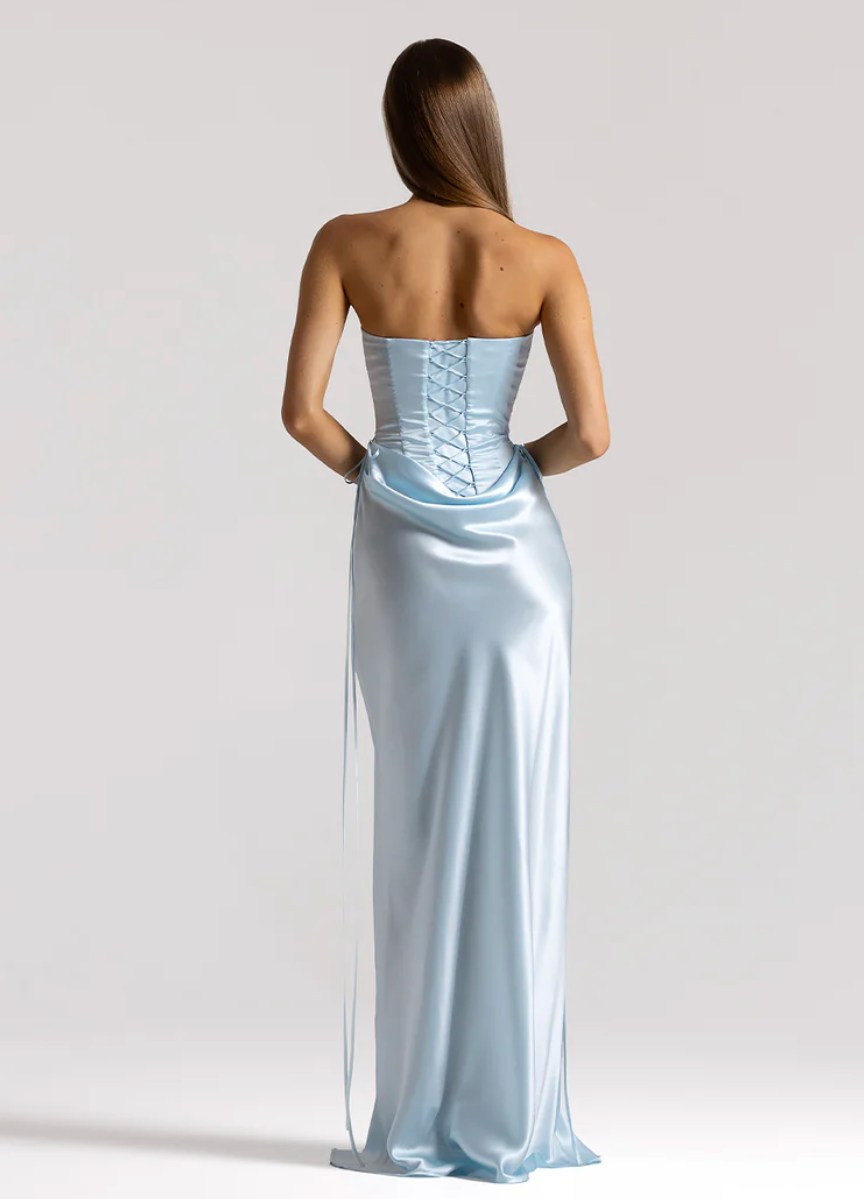 A woman stands with her back to the camera wearing a strapless light blue satin gown with a lace-up corset-style back and long side drapes, photographed against a plain light gray studio background.