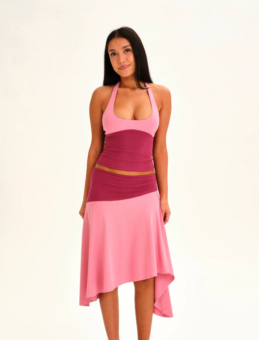 A woman modeling a color-blocked halter midi dress with a pink upper section, a darker magenta waistband, and a light pink flared skirt, set against a plain white studio background.