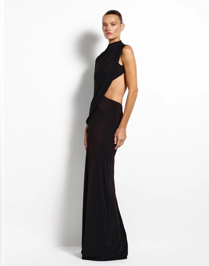 A model wearing a floor-length black gown with a high mock neck, draped shoulder detail, and a side cutout, photographed against a plain white studio background.