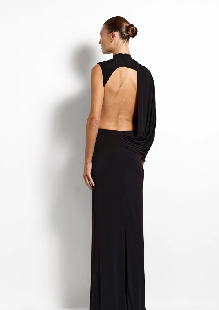 A model shown from the back wearing a black sleeveless maxi dress with a high neck, dramatic draped shoulder detail, and a high-waisted silhouette, set against a plain white studio background.