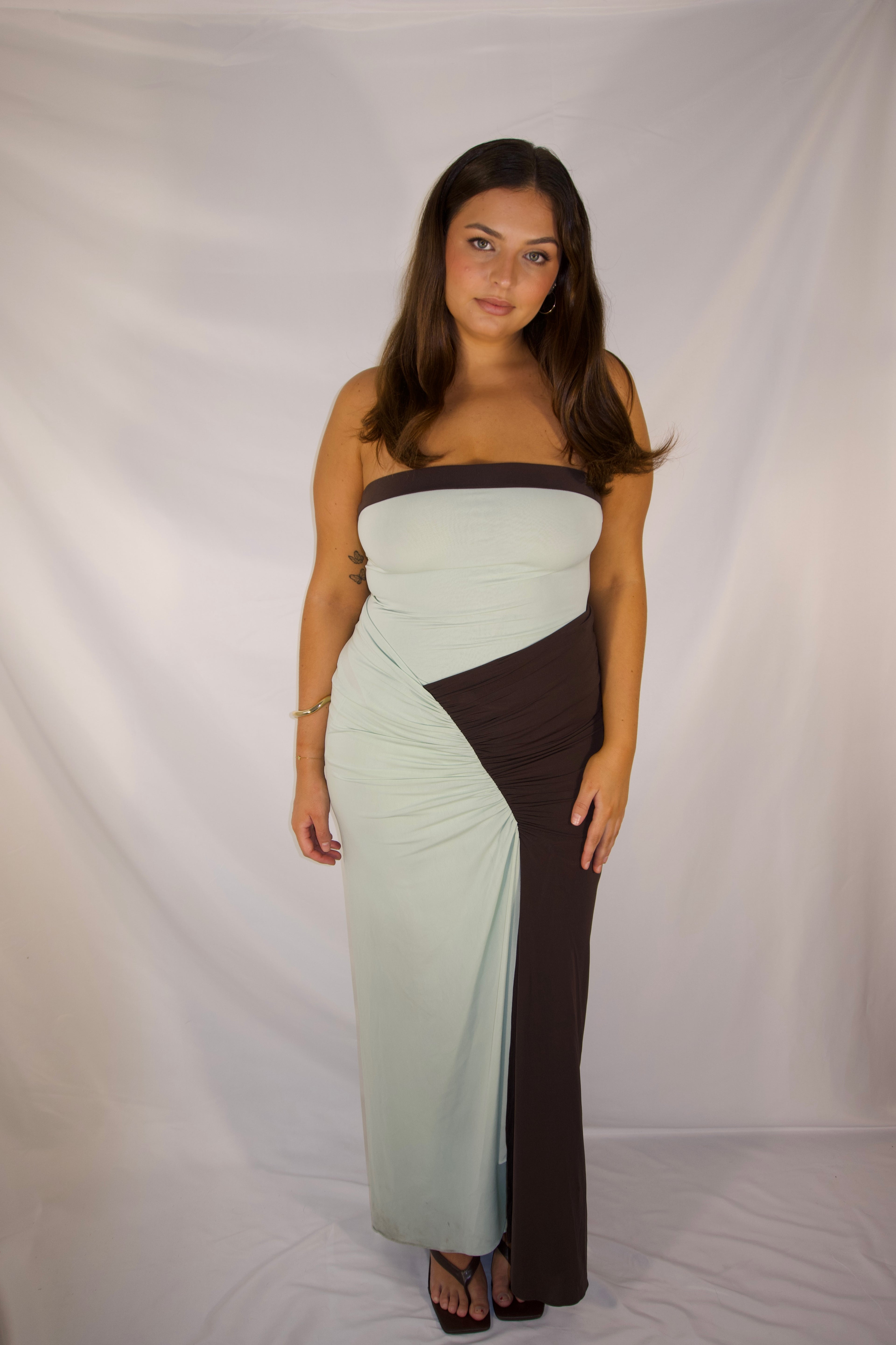 A woman modeling a strapless maxi dress with a color-blocked design in pale mint green and dark brown, standing against a white fabric backdrop.