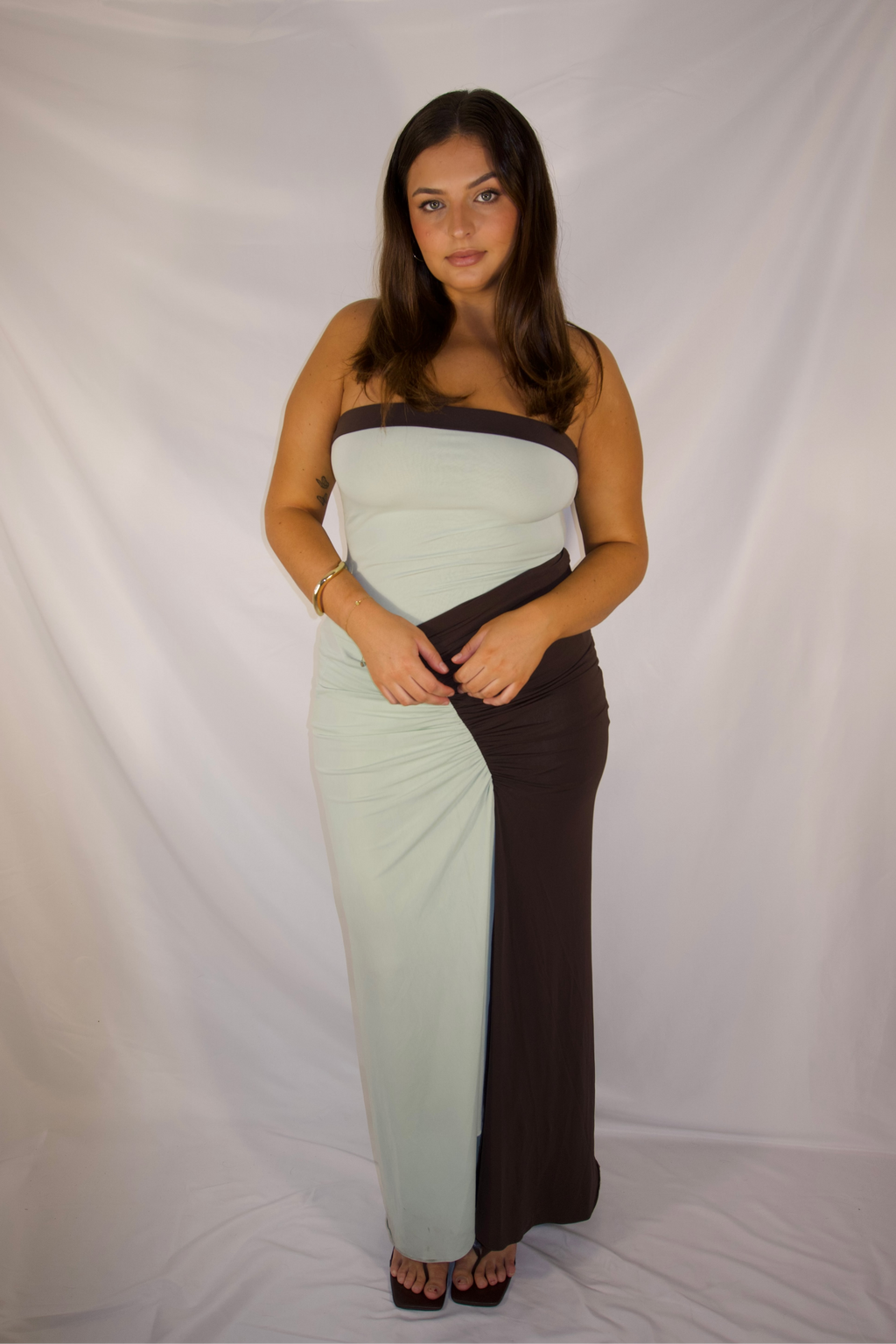 A woman modeling a strapless maxi dress with a color-blocked design in pale mint green and dark brown, standing against a white fabric backdrop.