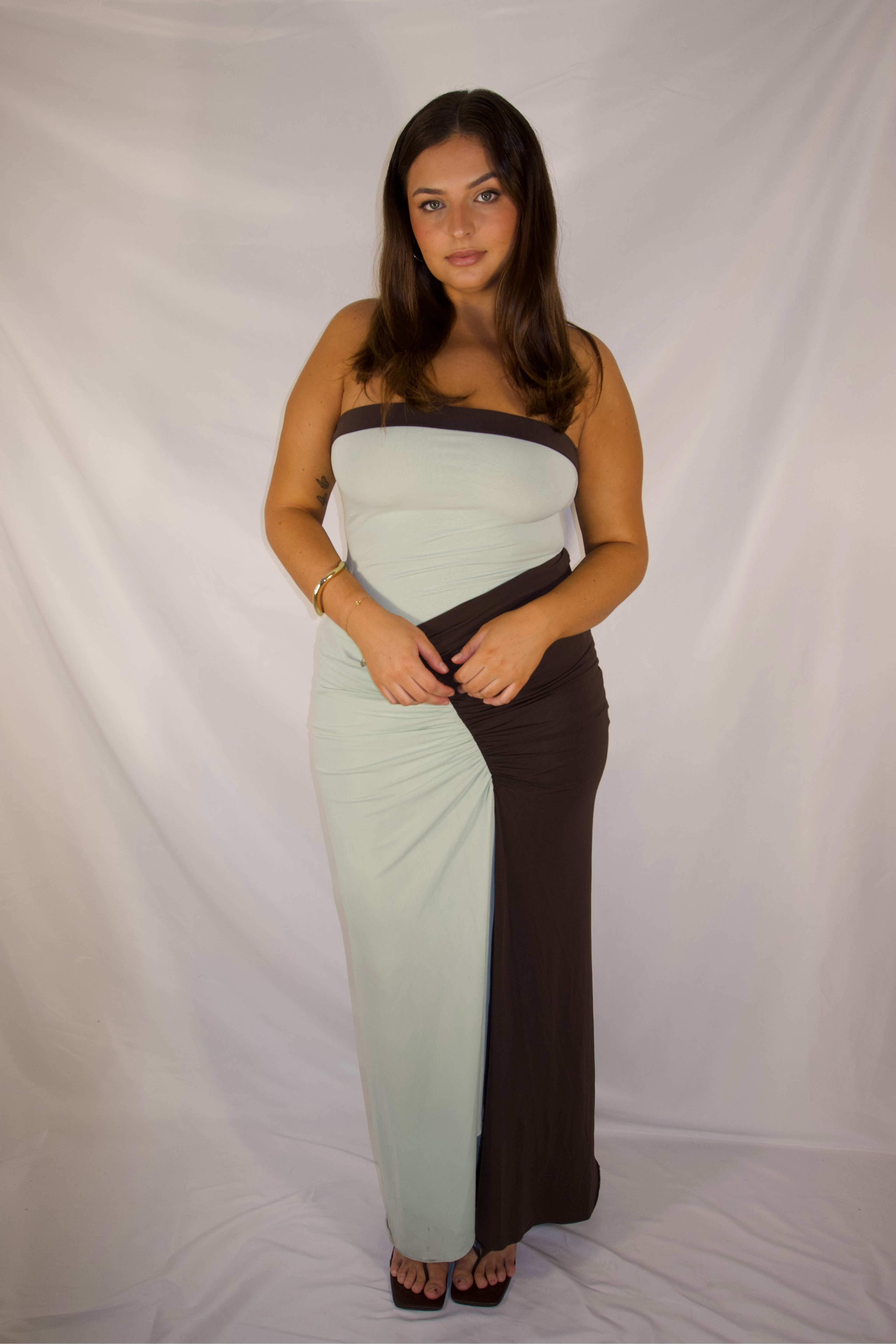 A woman modeling a strapless maxi dress with a color-blocked design in pale mint green and dark brown, standing against a white fabric backdrop.