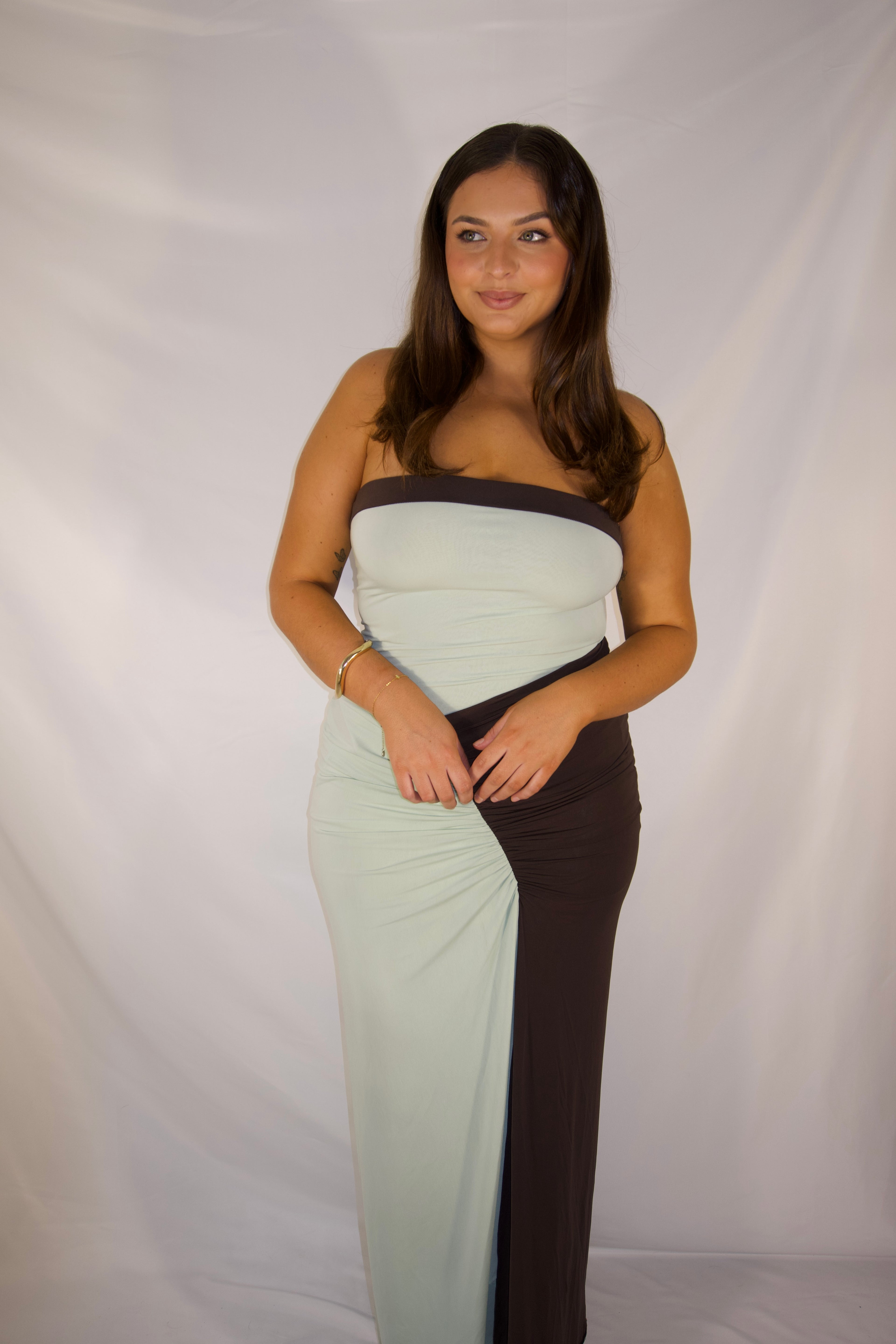 A woman modeling a strapless maxi dress with a color-blocked design in pale mint green and dark brown, standing against a white fabric backdrop.