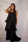 A woman modeling a black sleeveless tiered maxi dress with a draped, asymmetrical ruffle design, standing against a white fabric backdrop.