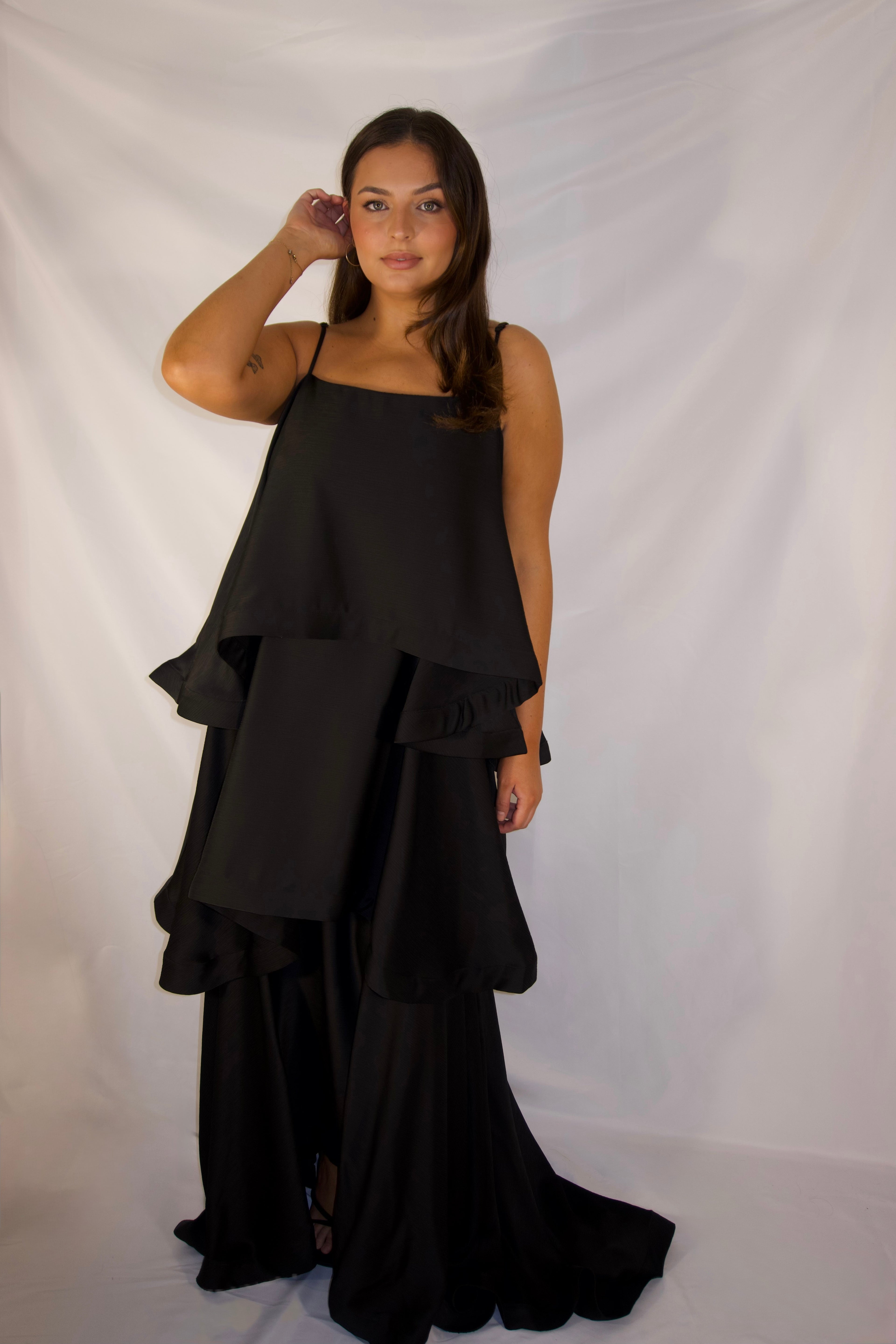 A woman modeling a black sleeveless tiered maxi dress with a draped, asymmetrical ruffle design, standing against a white fabric backdrop.