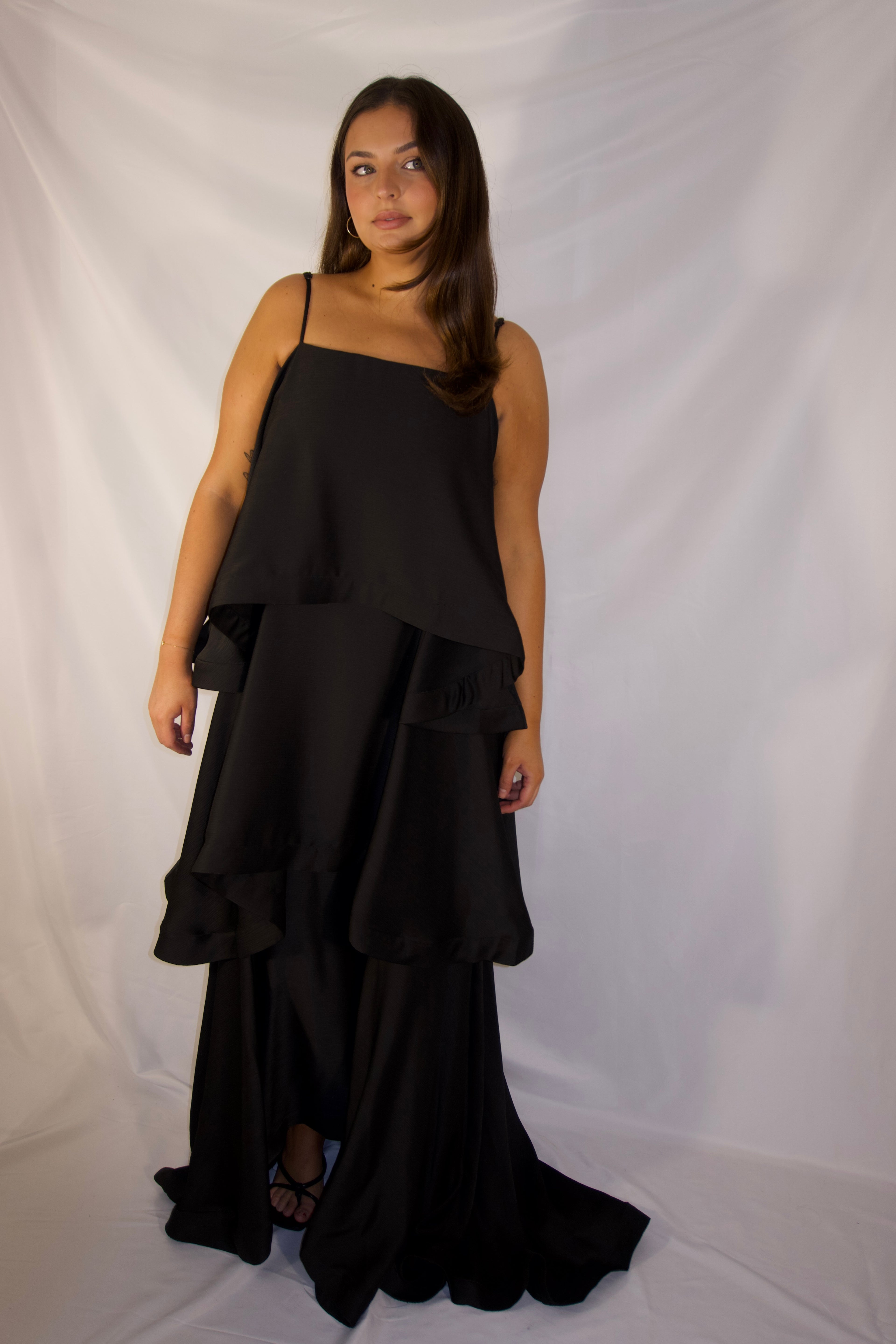 A woman modeling a black spaghetti-strap maxi dress with a layered, tiered ruffle skirt, standing against a white fabric backdrop.