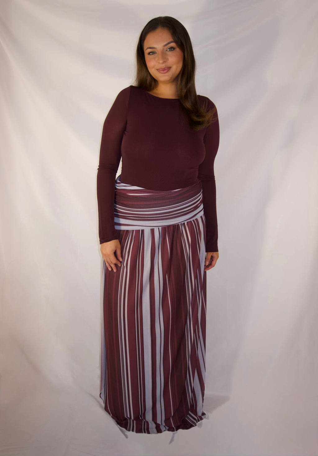 A woman modeling a two-piece outfit consisting of a long-sleeve maroon top and a matching long pleated skirt with vertical stripes in maroon, white, and gray, set against a draped white fabric backdrop.