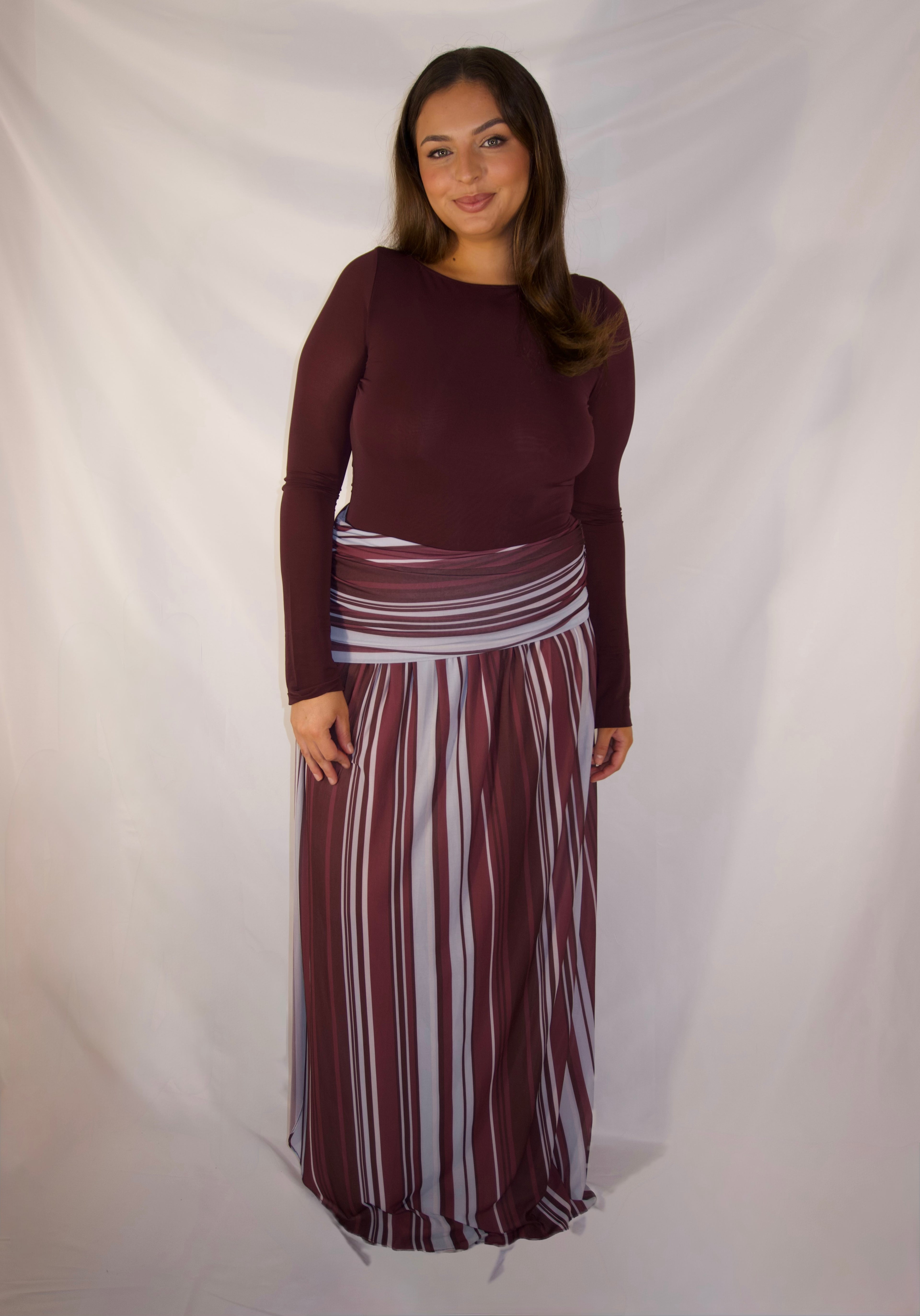 A woman modeling a two-piece outfit consisting of a long-sleeve maroon top and a matching long pleated skirt with vertical stripes in maroon, white, and gray, set against a draped white fabric backdrop.