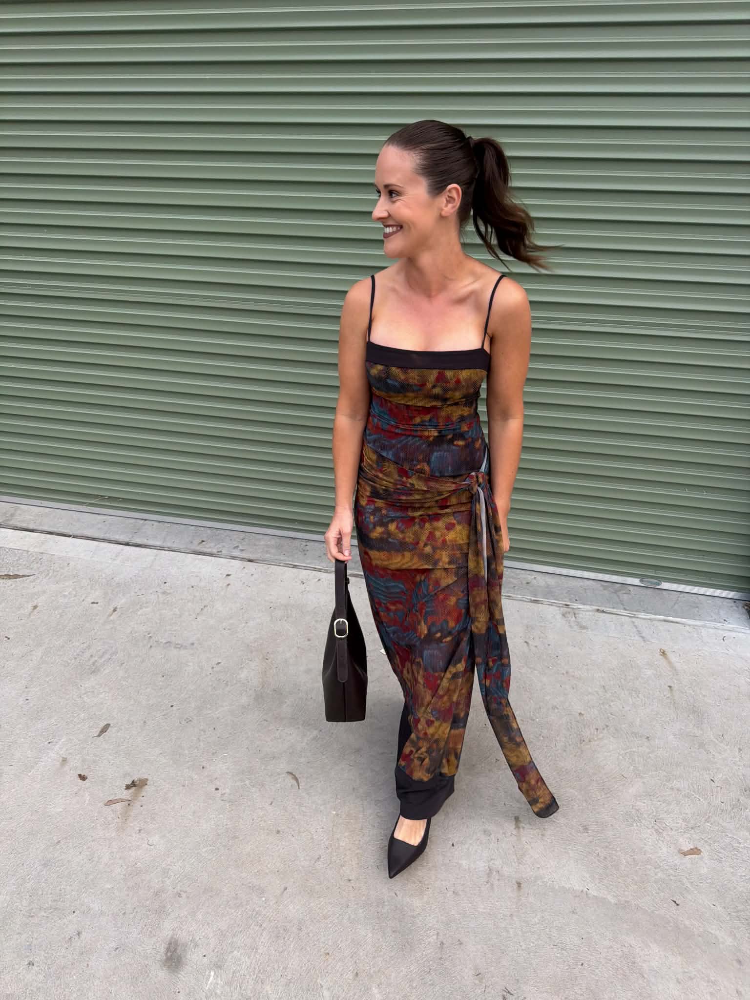 A woman wearing a sleeveless, multi-colored abstract print jumpsuit with thin straps and a side tie, standing in front of a green corrugated metal shutter.