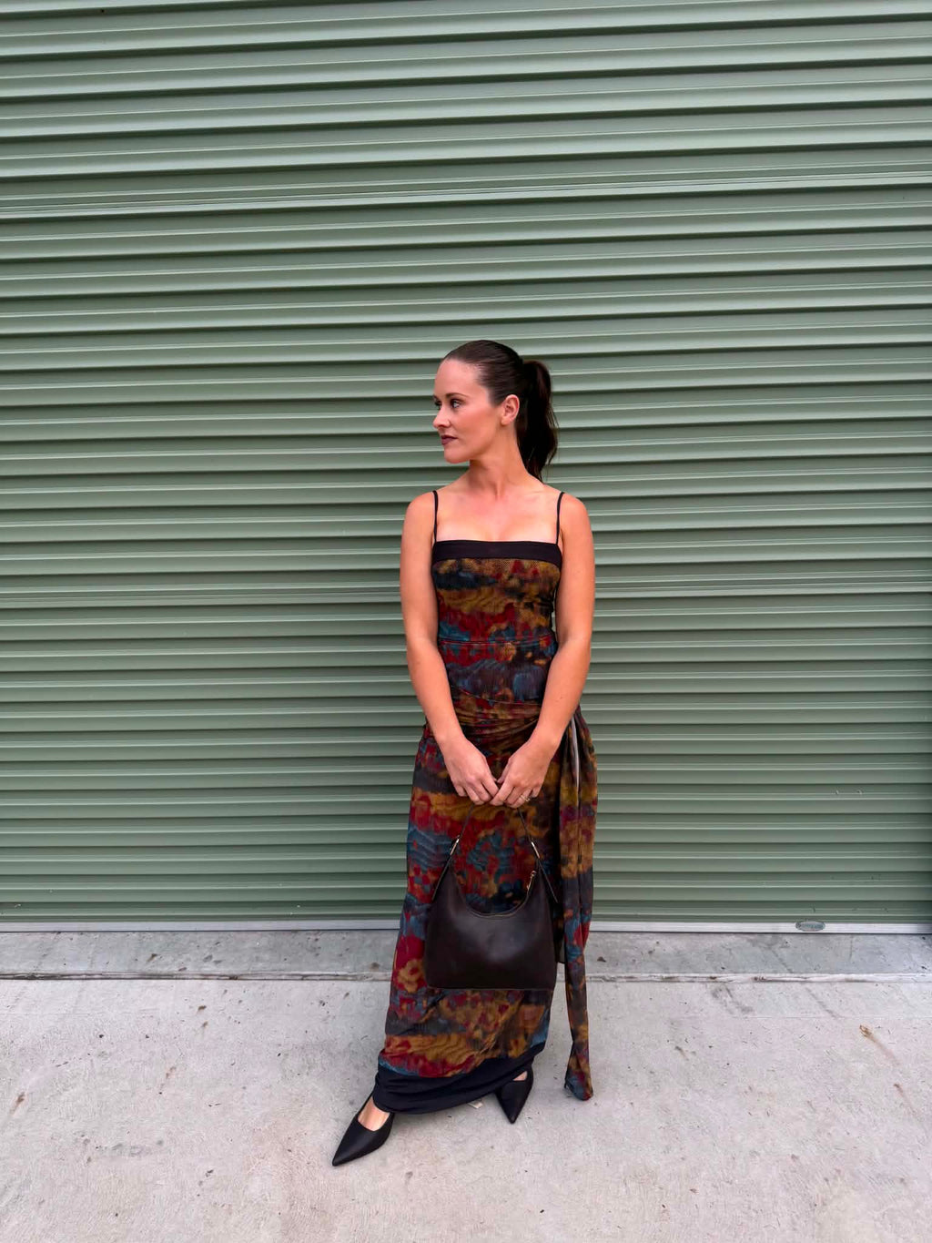 A woman stands in front of a green metal roll-up door wearing a long strapless maxi dress with a dark base and a colorful abstract pattern in red, blue, yellow, and brown. She is holding a dark brown handbag and wearing black pointed-toe flats.