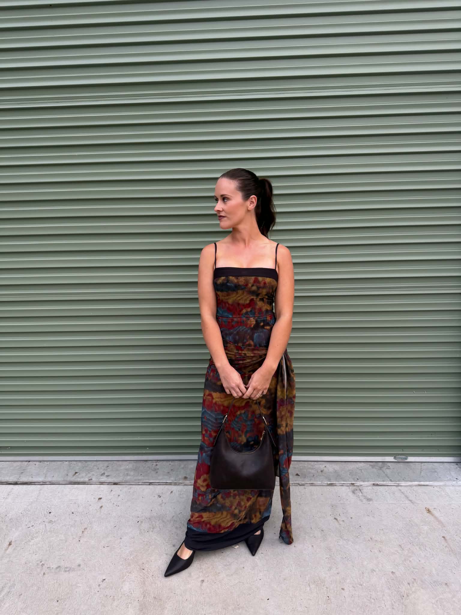 A woman stands in front of a green metal roll-up door wearing a long strapless maxi dress with a dark base and a colorful abstract pattern in red, blue, yellow, and brown. She is holding a dark brown handbag and wearing black pointed-toe flats.