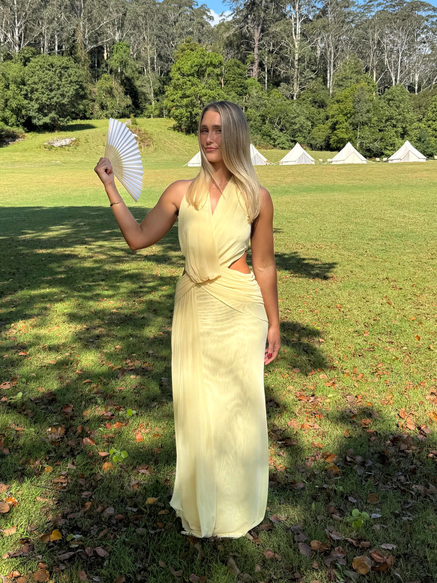 A woman stands in a grassy outdoor field holding a white folding fan, wearing a pale yellow sleeveless maxi dress. Trees, hills, and a blue sky are visible in the background, along with several white teepees.