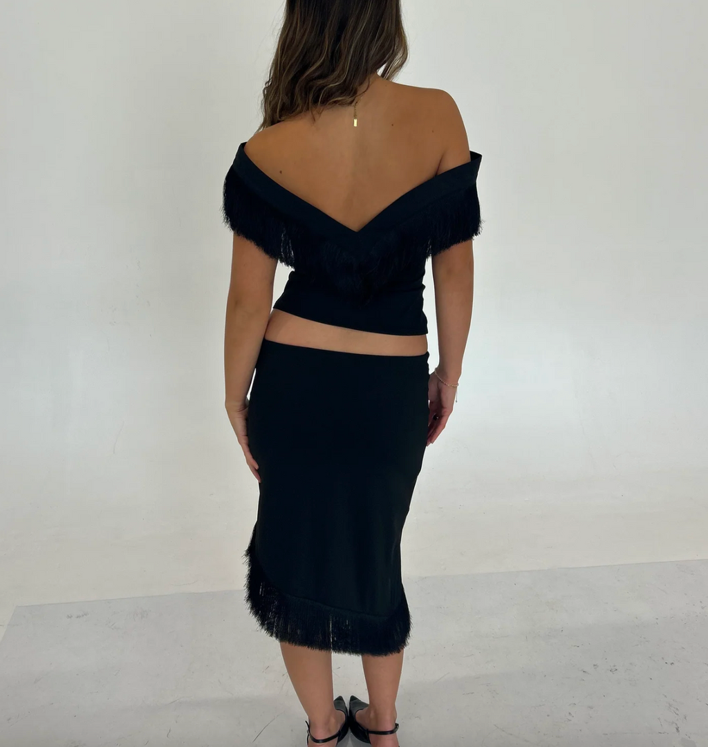 Back view of the Nalissa Luna Set in black, featuring an off-the-shoulder V-neck top with fringe detailing and a fitted, low-rise midaxi skirt with a fringed hem.